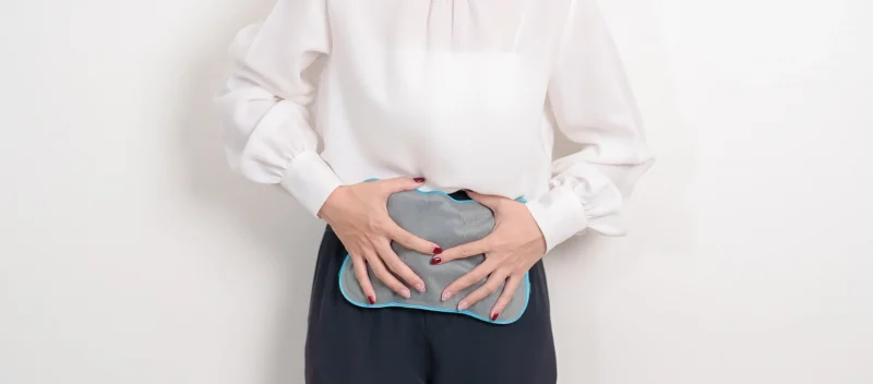 ภาพผู้หญิงใส่เสื้อสีขาวกำลังดูแลตัวเองเบื้องต้น เพื่อบรรเทาอาการปวดท้องบิดเกร็ง 